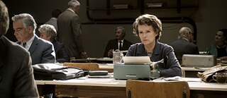 Colour image of an older white woman behind a table watching something outside the picture frame.