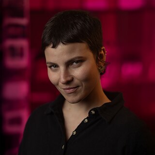 A foto mostra Martina di Gennaro diante de um fundo rosa, olhando diretamente para a câmara.