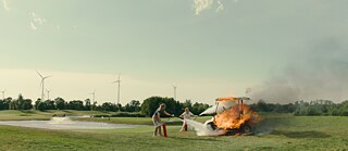 © Nikolaus GeyrhalterZwei Personen löschen ein brennendes Golfcart auf einer Wiese mit Feuerlöschern; im Hintergrund Windrad und Bäume.