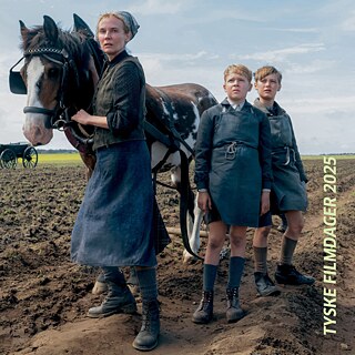 Drei Personen in Arbeitskleidung stehen auf einem Acker neben einem gescheckten Pferd; im Hintergrund weite Landschaft.