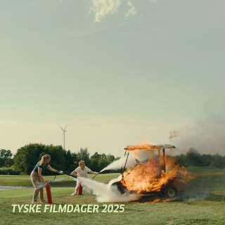 © Nikolaus Geyrhalter Filmproduktion Zwei Personen löschen ein brennendes Golfcart auf einer Wiese mit Feuerlöschern; im Hintergrund Windrad und Bäume.