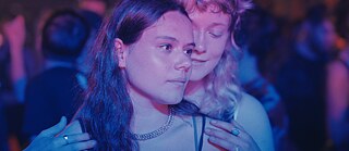 Zwei junge Frauen stehen eng beieinander in einer Umgebung mit bunten Lichtern, vermutlich auf einer Party oder Tanzfläche.