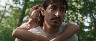 Zwei Personen im Freien, die Frau umarmt den Mann von hinten; im Hintergrund grüne Bäume.