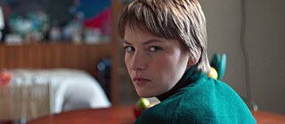 Person mit kurzem Haar in grünem Pullover sitzt mit dem Rücken zur Kamera, schaut aber direkt in die Kamera.