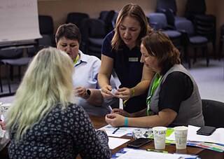 Учасники сидять застолом і тьторка зопомагає їм з виконанням завдання