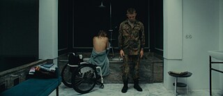 Eine Person in Militäruniform steht vor einer großen Badewanne, in der eine andere Person mit nacktem Rücken sitzt; daneben ein Rollstuhl.