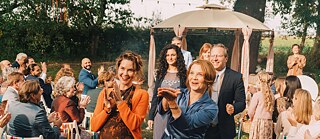 Gruppe von Menschen bei einer Feier im Freien, mit Stühlen, Dekoration und einem Pavillon im Hintergrund.
