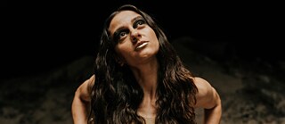 Person mit langen, gewellten Haaren steht in einer dunklen Umgebung und guckt nach oben, trägt ein ärmelloses Oberteil.