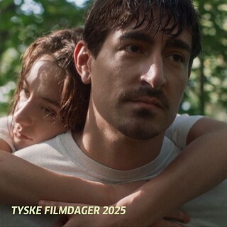 Zwei Personen im Freien, die Frau umarmt den Mann von hinten; im Hintergrund grüne Bäume.