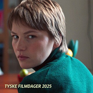 Person mit kurzem Haar in grünem Pullover sitzt mit dem Rücken zur Kamera, schaut aber direkt in die Kamera.