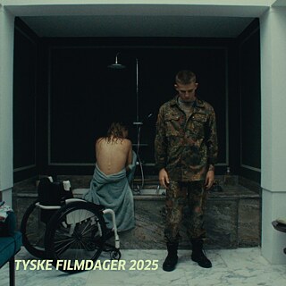 Eine Person in Militäruniform steht vor einer großen Badewanne, in der eine andere Person mit nacktem Rücken sitzt; daneben ein Rollstuhl.