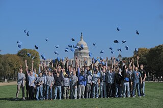 Children standing in a meadow throwing their light blue caps into the air, with the U.S. Capitol in Washington, D.C. in the background.
