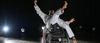 Ein Tänzer in einem Rollstuhl führt eine dynamische Tanzbewegung auf einer Bühne aus. Der Tänzer trägt ein graues Outfit und streckt die Arme und Beine in verschiedene Richtungen. Im Hintergrund sind mehrere Scheinwerfer zu sehen, die den dunklen Bühnenraum beleuchten.