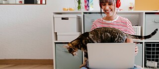 Eine junge Frau mit Laptop und rotem Kopfhörer lächelt, während eine Katze über die Tastatur geht