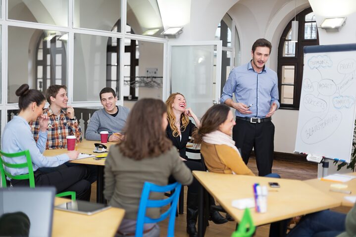 Ein Lehrer steht im Klassenraum vor einem Diagramm und erklärt etwas. Mehrere Studenten sitzen an Tischen und lachen herzlich.