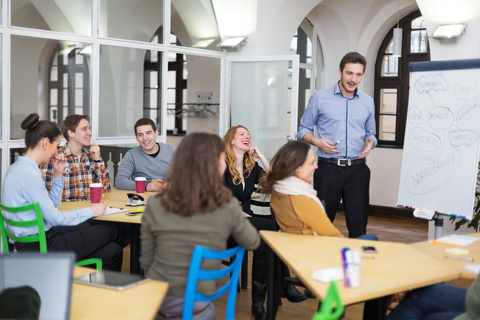 Teaser Fit-Formattraining Ein Lehrer steht im Klassenraum vor einem Diagramm und erklärt etwas. Mehrere Studenten sitzen an Tischen und lachen herzlich.