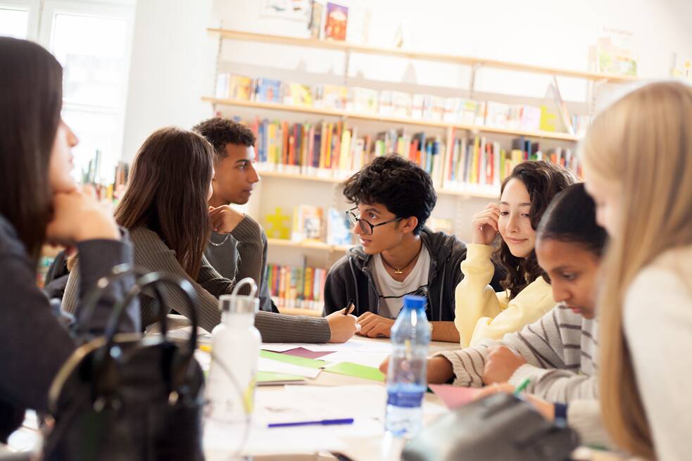 Eine Gruppe von Jugendlichen Camp Teilnehmenden sitzt und arbeitet an einem Tisch, auf welchem sich Zettel, Stifte und Flaschen befinden. Im Hintergrund sind Bücherregale zu sehen. Das Foto zeigt eine Gruppenarbeit im Deutschtraining. 