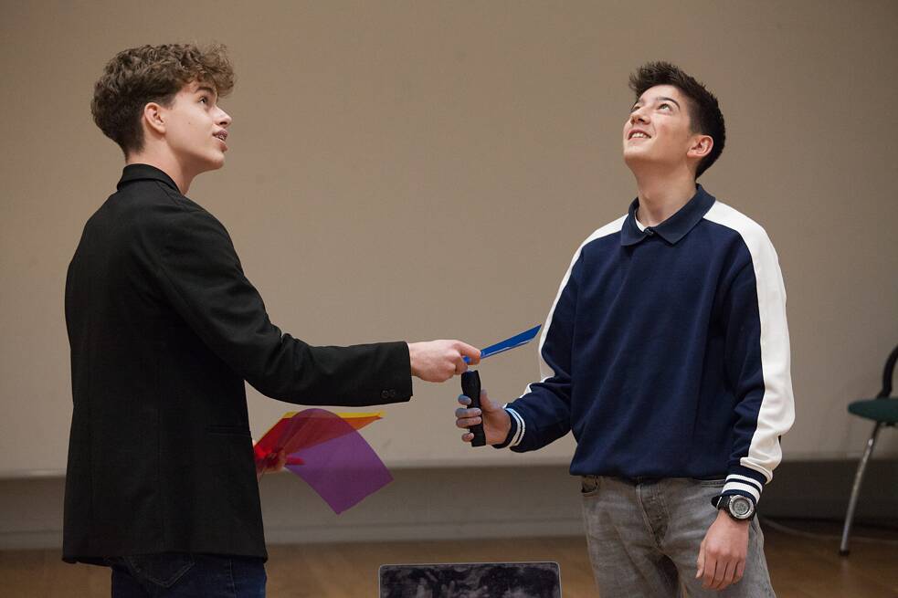 Zwei junge Männer stehen voreinander und schauen Richtung Decke. Der eine hält bunte Folien in seiner rechten Hand, der andere hält eine Taschenlampe darunter. 