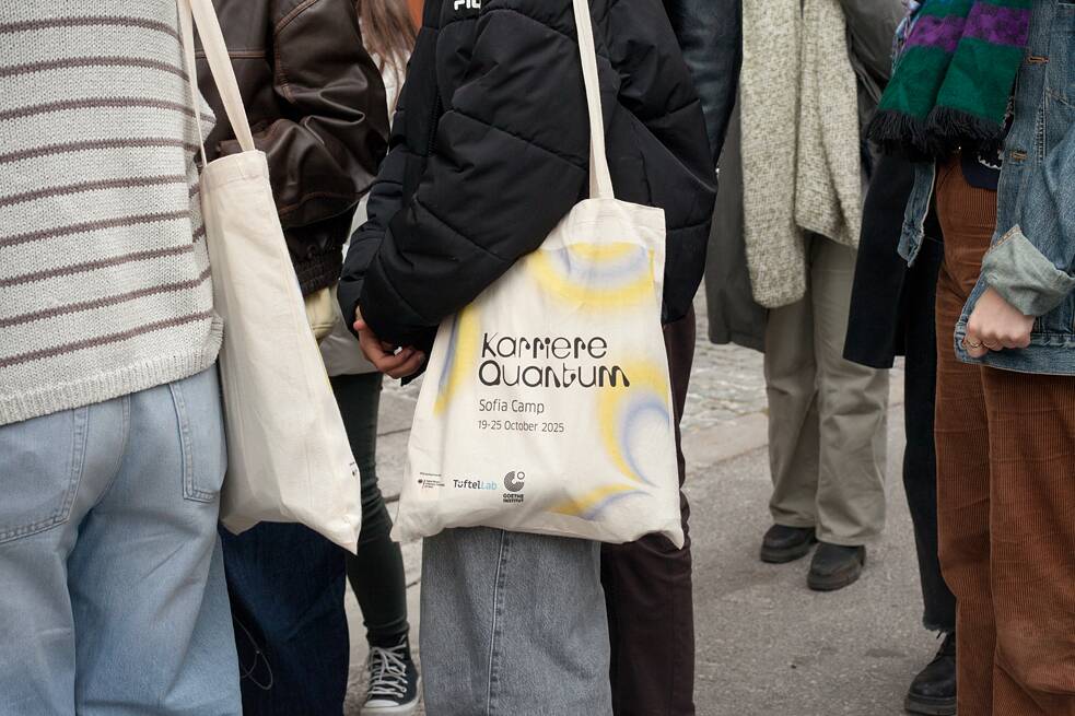 Mehrere Personen stehen zusammen im Freien, ein Mädchen mittig im Bild trägt eine Stofftasche mit der Aufschrift „Karriere Quantum – Sofia Camp, 19–25 October 2025“. 