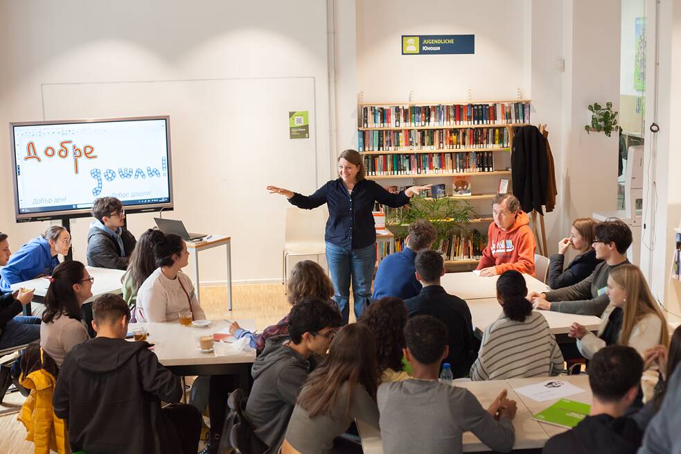 Eine Gruppe junger Menschen sitzt in einem Seminarraum und hört einer Vortragenden zu, die vor ihnen steht und gestikulierend spricht. Auf einem Bildschirm neben ihr steht auf bulgarisch „Willkommen!“. Im Hintergrund sind Bücherregale zu sehen. Das Foto zeigt die Bulgarisch-Stunde des Karriere-Quantum-Programms. 