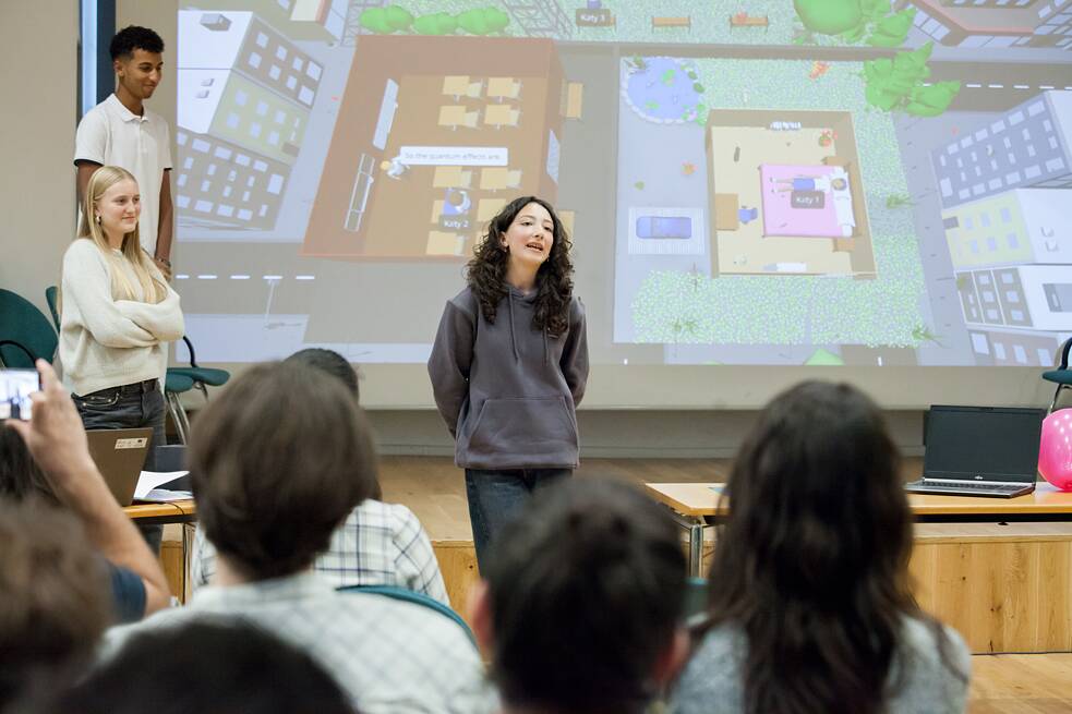 Drei Schülerinnen und Schüler stehen vor einer Leinwand, auf welcher eine bunte Projektion von Häusern aus der Vogelperspektive zu sehen ist. Menschen im Publikum sind von hinten zu sehen. 