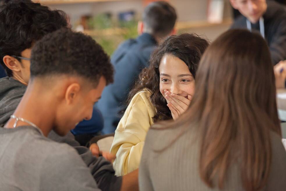 Auf dem Bild sind vier Personen zu sehen, zwei davon von hinten, einer von der Seite und ein Mädchen von vorn. Das Mädchen lacht und hält sich die Hände vor den Mund. Im Hintergrund sind weitere jugendliche Personen und Regale zu erkennen. 