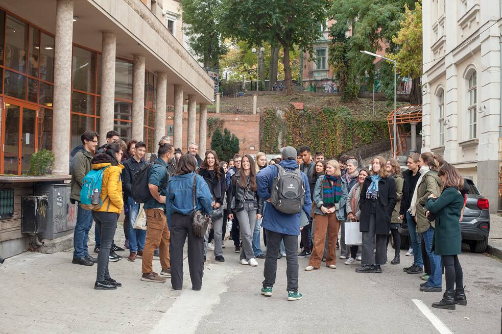 Eine große Gruppesteht in einer Seitenstraße, rechts und links befinden sich Gebäude. Ein Tour-Guide der Graffiti-Tour steht mit dem Rücken zur Gruppe, die Teilnehmenden hören ihm zu. 