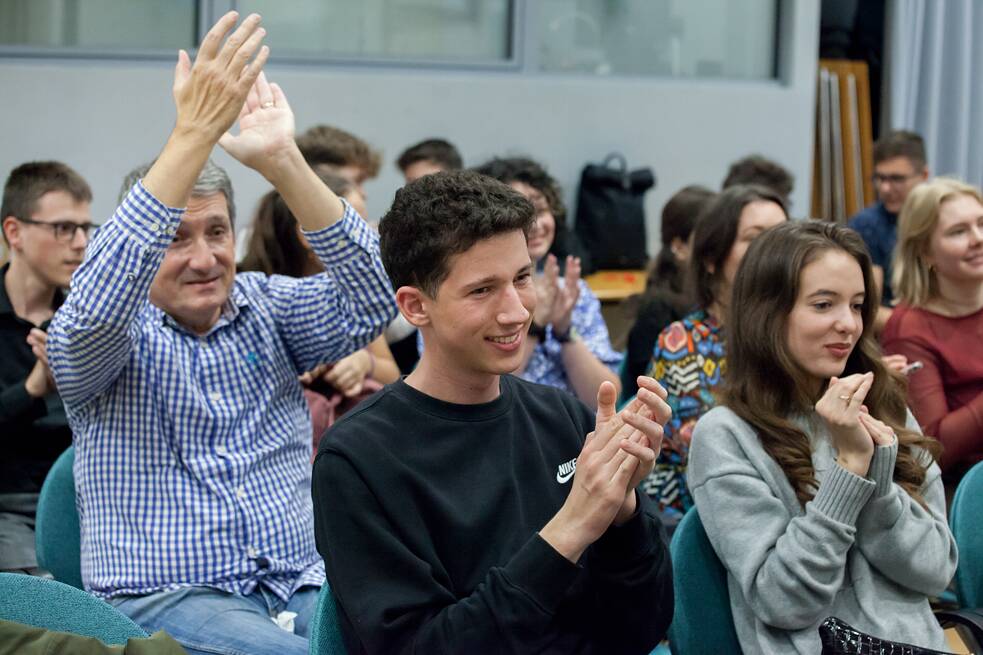 Applaudierende Schülerinnen und Schüler sowie ein älterer Mann sitzen im Publikum der Abschlussveranstaltung. Der ältere Mann applaudiert mit den Händen über dem Kopf. 
