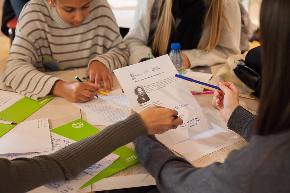 Vier Jugendliche sitzen an einem Tisch, auf welchem grüne Mappen und Zettel liegen. Sie haben Stifte in den Händen. Ein Mädchen hält ein Arbeitsblatt in der Hand, auf welchem ein Foto einer Person, gedruckter Text und handschriftliche Notizen zu sehen sind. Die Person neben ihr zeigt auf dieses Arbeitsblatt. Das Foto zeigt eine Deutsch-Unterrichtsstunde. 