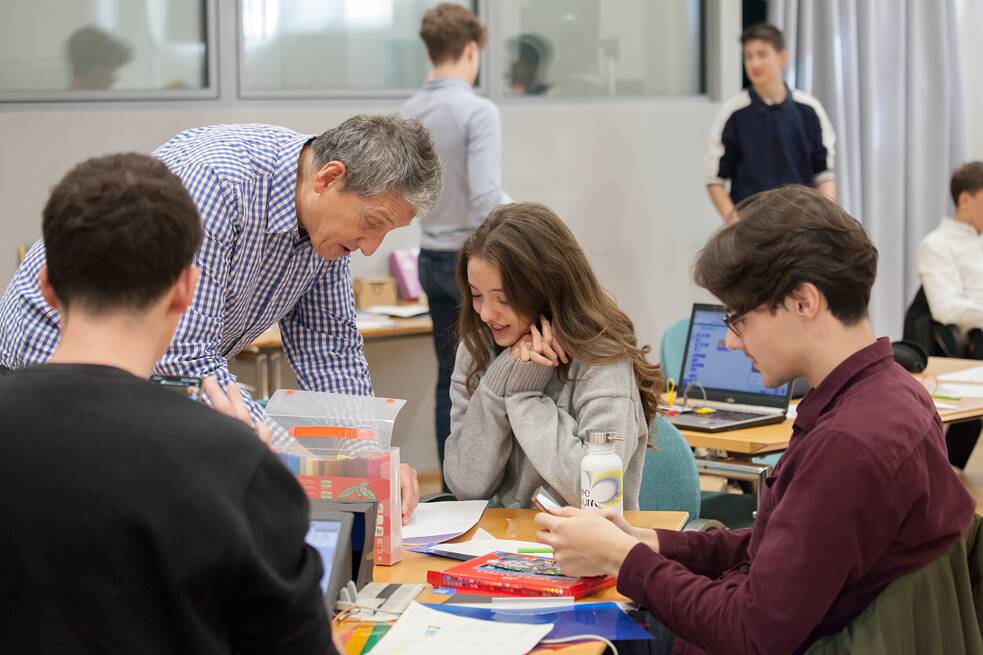 Vier Personen, davon zwei Schüler, eine Schülerin und ein älterer Lehrer sind um einen Tisch versammelt. Die Schülerinnen und Schüler sitzen, zwei von ihnen bedienen ein Handy. Der Lehrer steht und zeigt etwas der Schülerin, deren Blick nach unten gerichtet ist. Im Hintergrund sind weitere stehende Personen, Tische und Laptops zu sehen. 