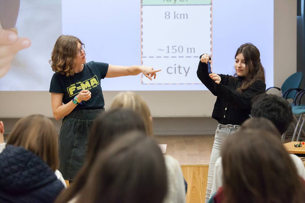 Zwei Personen stehen vor einer Präsentationsleinwand. Eine von ihnen zeigt auf einen kleinen Gegenstand, den die andere in der Hand hält, während das Publikum ihnen aufmerksam zuschaut.  