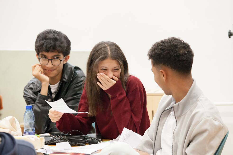 Auf dem Bild sind drei sitzende Teenager zu sehen. In der Mitte befindet sich ein Mädchen, welches ein weißes Blatt Papier in der Hand hält und lachend die Hand vor den Mund hält. Die Jungen rechts und links neben ihr schauen in ihre Richtung. Auf dem Tisch vor ihnen steht eine Flasche und es liegen ein Kabel und weitere Zettel darauf. 