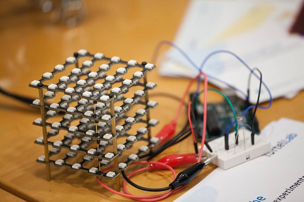 Ein LED-Würfel aus mehreren Schichten kleiner LEDs ist mit Kabeln und einem Breadboard verbunden. Im Hintergrund liegen Unterlagen und eine Mikrocontroller-Platine auf einem Tisch. Das Foto entstand im Rahmen eines TüftelLab-Workshops. 