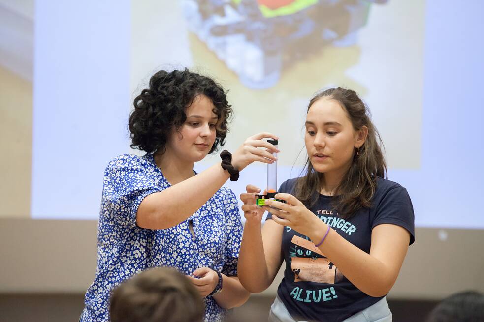 Zwei Jugendliche stehen gemeinsam vor einer Leinwand und präsentieren ein kleines technisches Objekt, das sie in den Händen halten. Das Foto entstand bei der Abschlussveranstaltung des Camps. 
