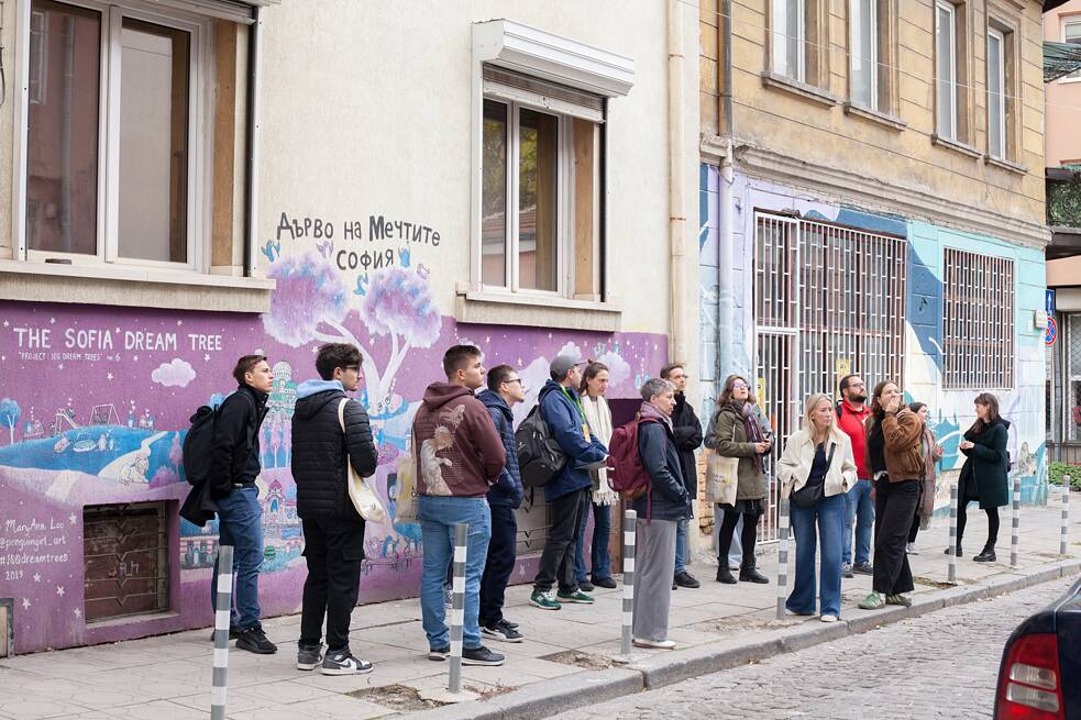 14 Teilnehmende des Camps und Mitglieder des Teams stehen auf dem Bürgersteig und schauen in eine Richtung. Hinter ihnen ist ein Graffiti an einer Hauswand zu sehen, auf welchem “The Sofia Dream Tree” steht. 