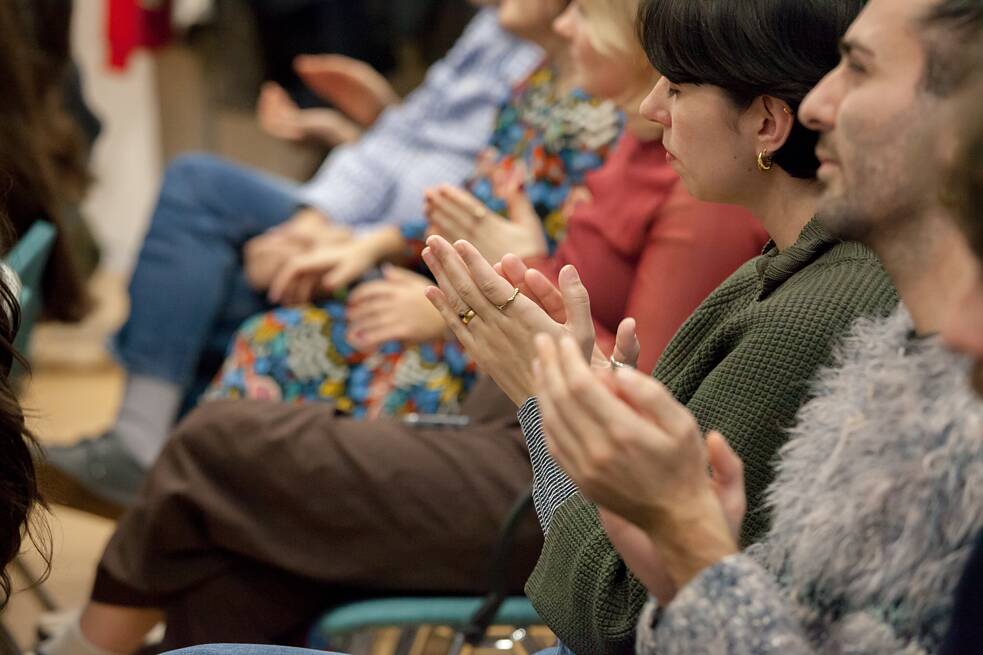 Es sind mehrere Personen von der Seite zu sehen, welche im Publikum der Abschlussveranstaltung sitzen und applaudieren.  