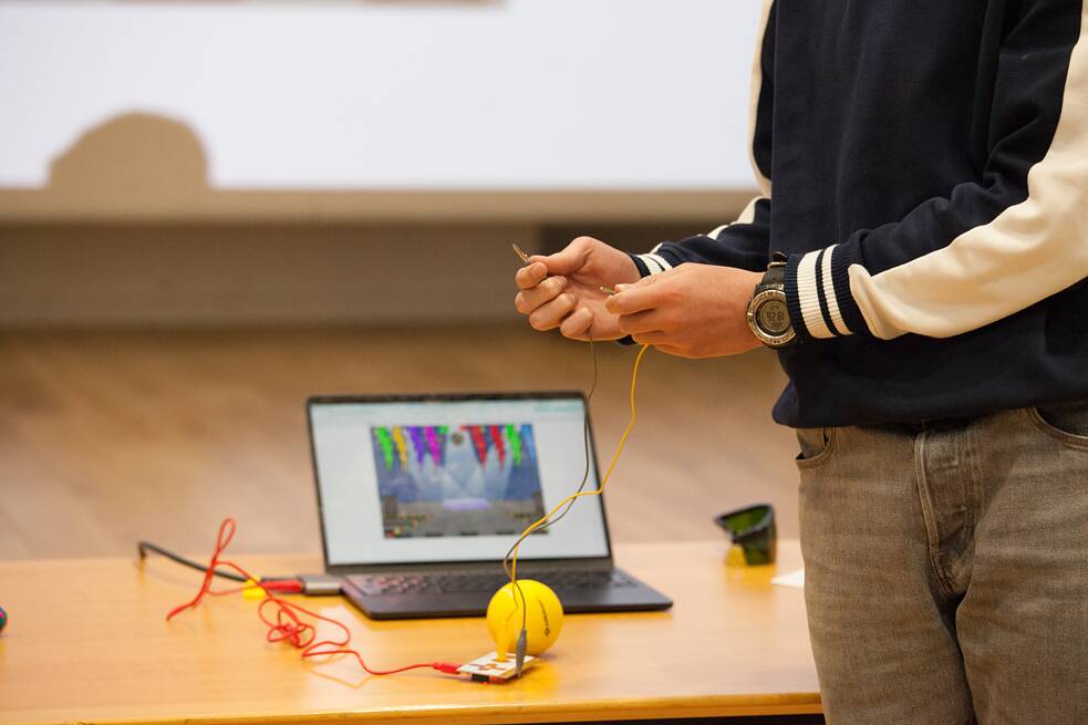 Auf dem Bild ist ein Campteilnehmer zu sehen, der zwei Kabel in der Hand hält. Diese Kabel sind an einem technischen Gerät angeschlossen, welches zusammen mit einem Laptop auf einem Tisch hinter der Person steht. 
