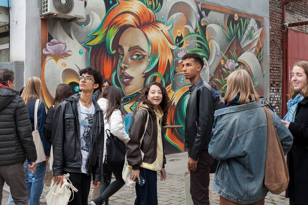 Eine Gruppe von Camp Teilnehmenden nimmt an einer Graffiti-Tour durch Sofia teil. Sie stehen vor einer großflächigen, farbenfrohen Wandmalerei mit einem Porträtmotiv und betrachten die Kunstwerke interessiert.  