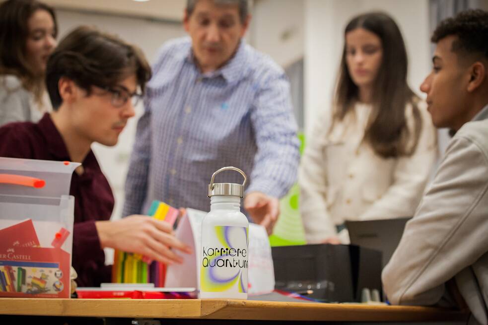 Zu sehen ist eine Flasche mit dem Aufdruck “Karriere Quantum”, welche auf einem Tisch steht. Dahinter sind vier Schülerinnen und Schüler sowie ein Lehrer zu sehen. 