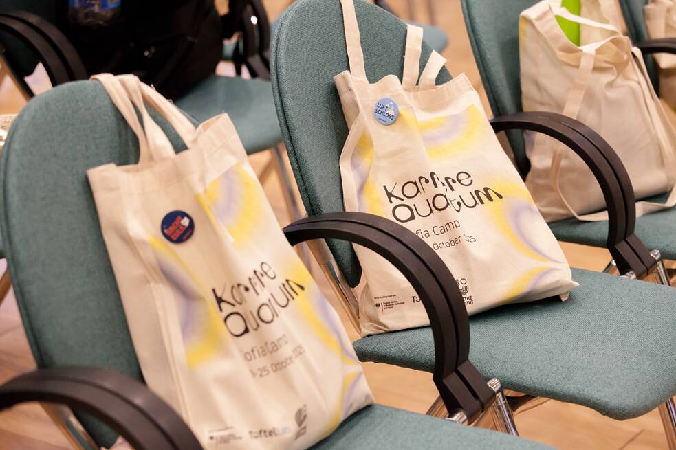 Drei Stühle stehen nebeneinander. Auf ihnen stehen Stoffbeutel mit dem Schriftzug und Logo des Karriere Quantum Camps. Auf den Beuteln sind zudem Buttons des Goethe-Instituts angebracht. 
