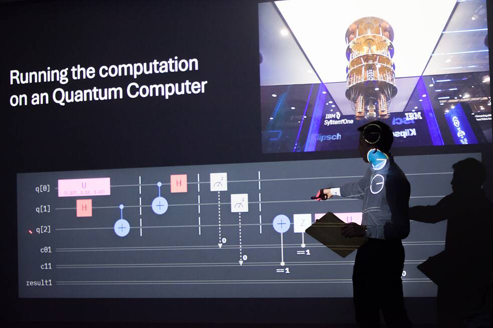 Eine Person steht vor einer Leinwandprojektion, auf welcher “Running the computation on a Quantum Computer” zu lesen ist. Die Person wird von der Präsentation beleuchtet und zeigt auf eine technische Abbildung unterhalb des Schriftzugs. 
