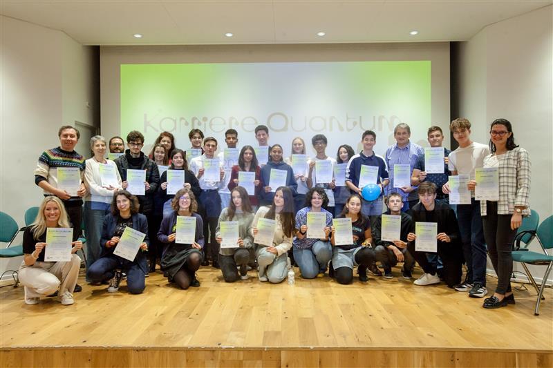 Eine große Gruppe jüngerer Menschen steht auf einer Bühne und hält Zertifikate in den Händen. Im Hintergrund ist eine Präsentationsfolie mit dem Schriftzug „Karriere Quantum“ zu sehen. Alle lächeln in die Kamera. 