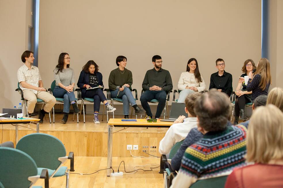 Neun Personen sitzen im Halbkreis auf einer Bühne. Eine Frau am Rand hält ein Mikrofon in der Hand und schaut zu den Personen, die neben ihr sitzen. Im Vordergrund sitzen Menschen im Publikum, zu sehen sind diese von hinten. 