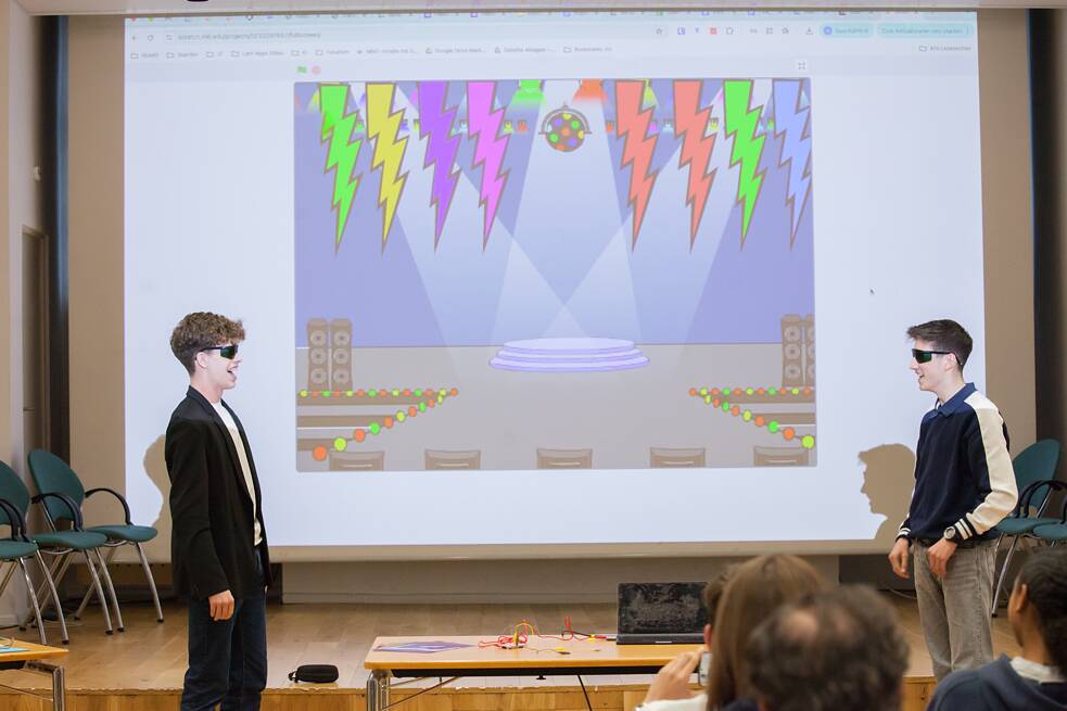 Zwei Schüler stehen vor einer großen Leinwand in einem Präsentationsraum und präsentieren ein bunt animiertes digitales Bühnenbild, das auf der Leinwand angezeigt wird. Auf der Bühne im Hintergrund sitzen mehrere Leute und beobachten die Präsentation. 
