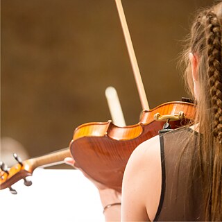 Das Foto zeigt eine Nahaufnahme von einer Person von hinten, wie sie ein Streichinstrument spielt. Man sieht einen Teil der linken Schult und Teil des Hinterkopfes.