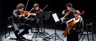 Das Bild zeigt vier Musiker*innen, die unterschiedliche Streichinstrumente spielen. Vor ihnen steht jeweils ein Notenständer mit Notenblättern.