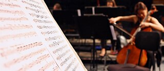 Zeigt auf der linken Seite in Nahaufnahme ein Notenblatt und rechts im Hintergrund unscharf fotografiert, eine Person, die sitzend ein Streichinstrument spielt.