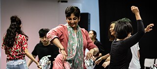 A group of people is dancing in a studio setting. One person in the foreground is wearing a pink outfit with a patterned scarf, while others around are moving energetically. Silver balloons are visible on the floor.