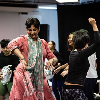 A group of people is dancing in a studio setting. One person in the foreground is wearing a pink outfit with a patterned scarf, while others around are moving energetically. Silver balloons are visible on the floor.