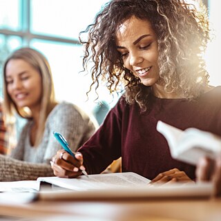 Zwei junge Frauen lesen lächelnd aus Büchern und Arbeitsblättern